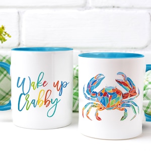 May include: Two white ceramic mugs with blue rims. One mug has the text "Wake up Crabby" in rainbow colors. The other mug has a colorful illustration of a crab.