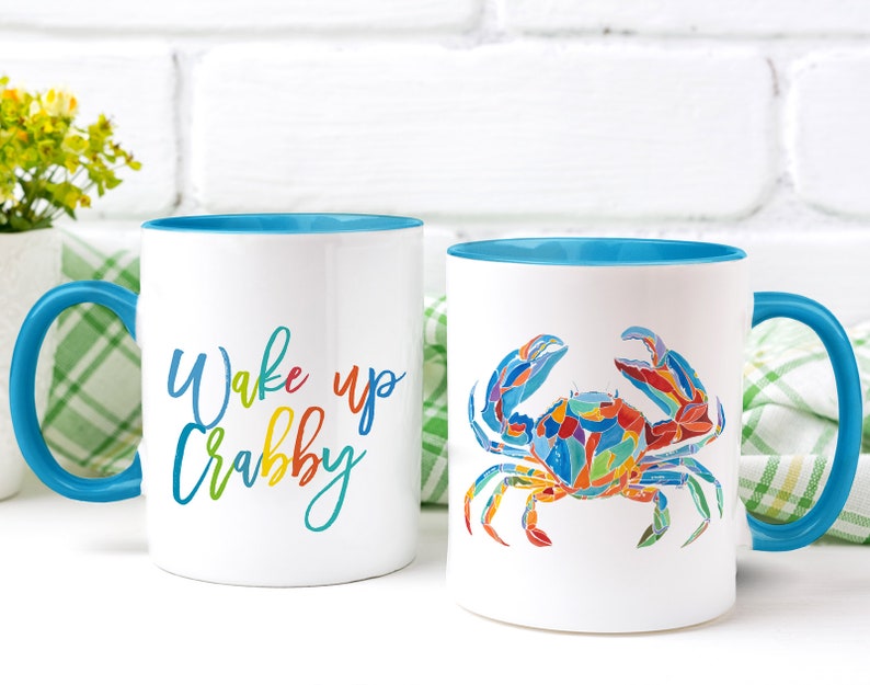 May include: Two white ceramic mugs with blue rims. One mug has the text "Wake up Crabby" in rainbow colors. The other mug has a colorful illustration of a crab.