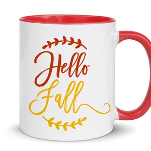 K&ouml;nnte beinhalten: Wei&szlig;e Keramiktasse mit rotem Rand und Henkel. Die Tasse hat ein Design mit der Aufschrift "Hello Fall" in roten und gelben Buchstaben mit einem Blattdesign.