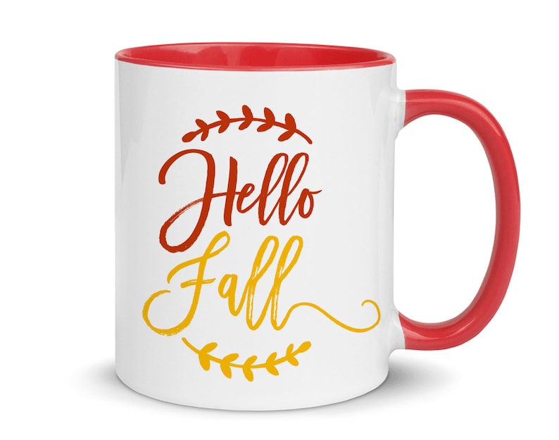 K&ouml;nnte beinhalten: Wei&szlig;e Keramiktasse mit rotem Rand und Henkel. Die Tasse hat ein Design mit der Aufschrift "Hello Fall" in roten und gelben Buchstaben mit einem Blattdesign.