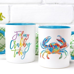 May include: Two white ceramic mugs with blue rims. One mug has the text "Feeling Crabby Today" in rainbow colors. The other mug has a colorful illustration of a crab.