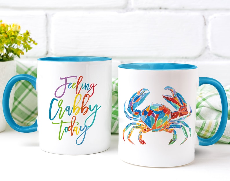 May include: Two white ceramic mugs with blue rims. One mug has the text "Feeling Crabby Today" in rainbow colors. The other mug has a colorful illustration of a crab.