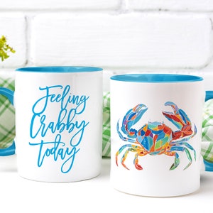 May include: Two white ceramic mugs with blue rims. One mug has the text "Feeling Crabby Today" in blue script. The other mug has a colorful illustration of a crab.