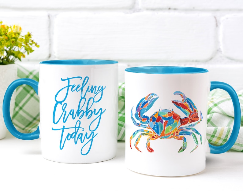 May include: Two white ceramic mugs with blue rims. One mug has the text "Feeling Crabby Today" in blue script. The other mug has a colorful illustration of a crab.