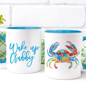 May include: Two white ceramic mugs with blue rims. One mug has the text "Wake up Crabby" in blue lettering. The other mug has a colorful illustration of a crab.