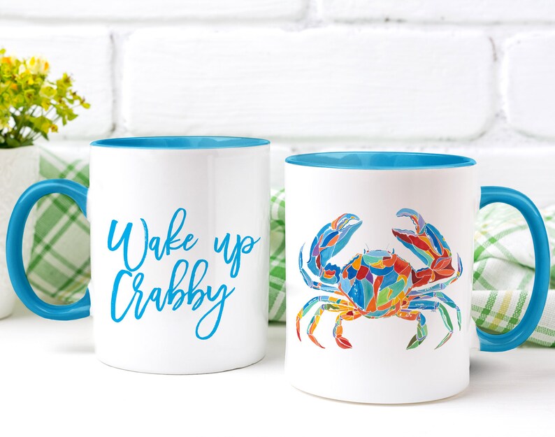 May include: Two white ceramic mugs with blue rims. One mug has the text "Wake up Crabby" in blue lettering. The other mug has a colorful illustration of a crab.