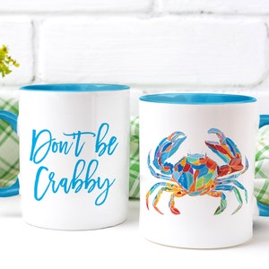 May include: Two white ceramic mugs with blue rims and handles. One mug has the text "Don't be Crabby" in blue lettering. The other mug has a colorful illustration of a crab.