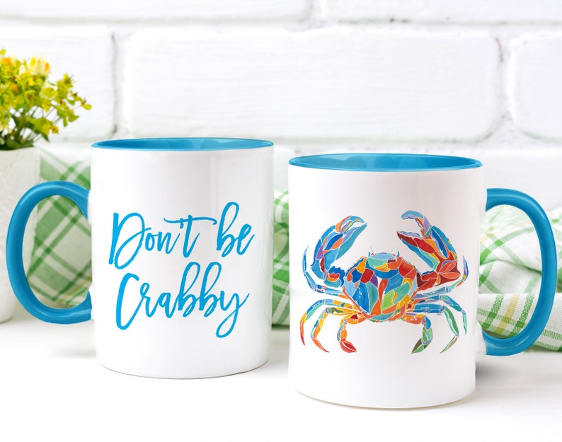 May include: Two white ceramic mugs with blue rims and handles. One mug has the text "Don't be Crabby" in blue lettering. The other mug has a colorful illustration of a crab.