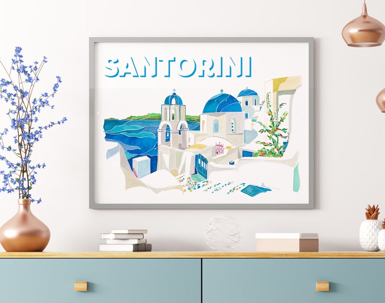 K&ouml;nnte beinhalten: Ein Aquarellbild eines wei&szlig;en Geb&auml;udes mit blauen Kuppeln in Santorini, Griechenland. Das Bild ist gerahmt und tr&auml;gt den Schriftzug "SANTORINI" in blauen Buchstaben oben.