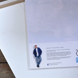 May include: A white card with a blue and white watercolour background. The card has text that reads "About the Artist, Jack" and includes a photo of the artist, Jack. The text describes Jack as a French Montrealer born in Brittany, France. The text also describes his paintings as being inspired by his life experiences, travels to Italy and the Caribbean, and his children's tales. The text states that the print is a reproduction of his original watercolour painting printed on professional acid-free quality paper using archival inks that will resist fading.