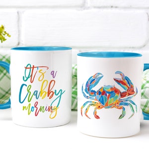 May include: Two white ceramic mugs with blue rims. One mug has the text "It's a Crabby Morning" in rainbow colors. The other mug has a colorful illustration of a crab.