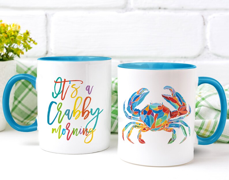May include: Two white ceramic mugs with blue rims. One mug has the text "It's a Crabby Morning" in rainbow colors. The other mug has a colorful illustration of a crab.