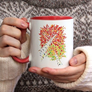 K&ouml;nnte beinhalten: Wei&szlig;e Keramiktasse mit rotem Rand, die eine farbenfrohe abstrakte Malerei eines Birkenbaums mit Herbstlaub zeigt. Die Tasse wird von einer Person gehalten, die einen wei&szlig;en Pullover tr&auml;gt.
