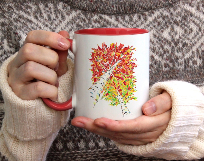 K&ouml;nnte beinhalten: Wei&szlig;e Keramiktasse mit rotem Rand, die eine farbenfrohe abstrakte Malerei eines Birkenbaums mit Herbstlaub zeigt. Die Tasse wird von einer Person gehalten, die einen wei&szlig;en Pullover tr&auml;gt.