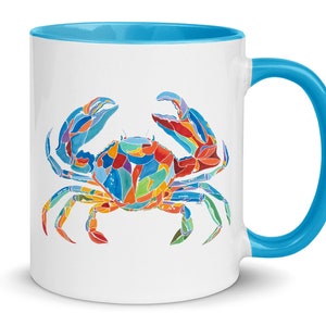 May include: A white ceramic mug with a blue rim and handle. The mug features a colorful illustration of a crab with blue, red, orange, yellow, and green accents.