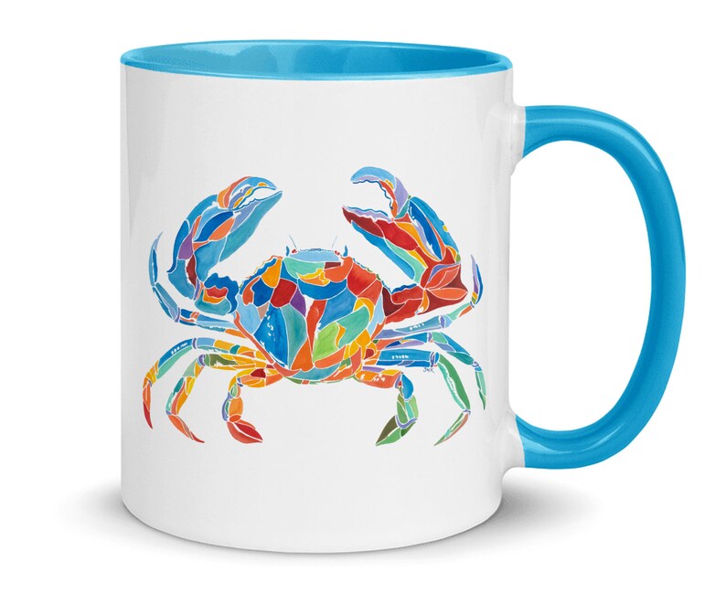 May include: A white ceramic mug with a blue rim and handle. The mug features a colorful illustration of a crab with blue, red, orange, yellow, and green accents.