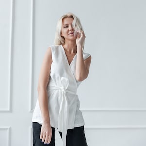 May include: A woman with blonde hair is wearing a white linen wrap top with a tie at the waist. She is standing in front of a white wall.