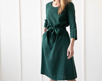 May include: A woman wearing a dark green linen dress with a self-tie belt. The dress has a round neckline and three-quarter sleeves.