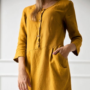 May include: A mustard yellow linen dress with a button-up front and pockets. The dress has a relaxed fit and a midi length.