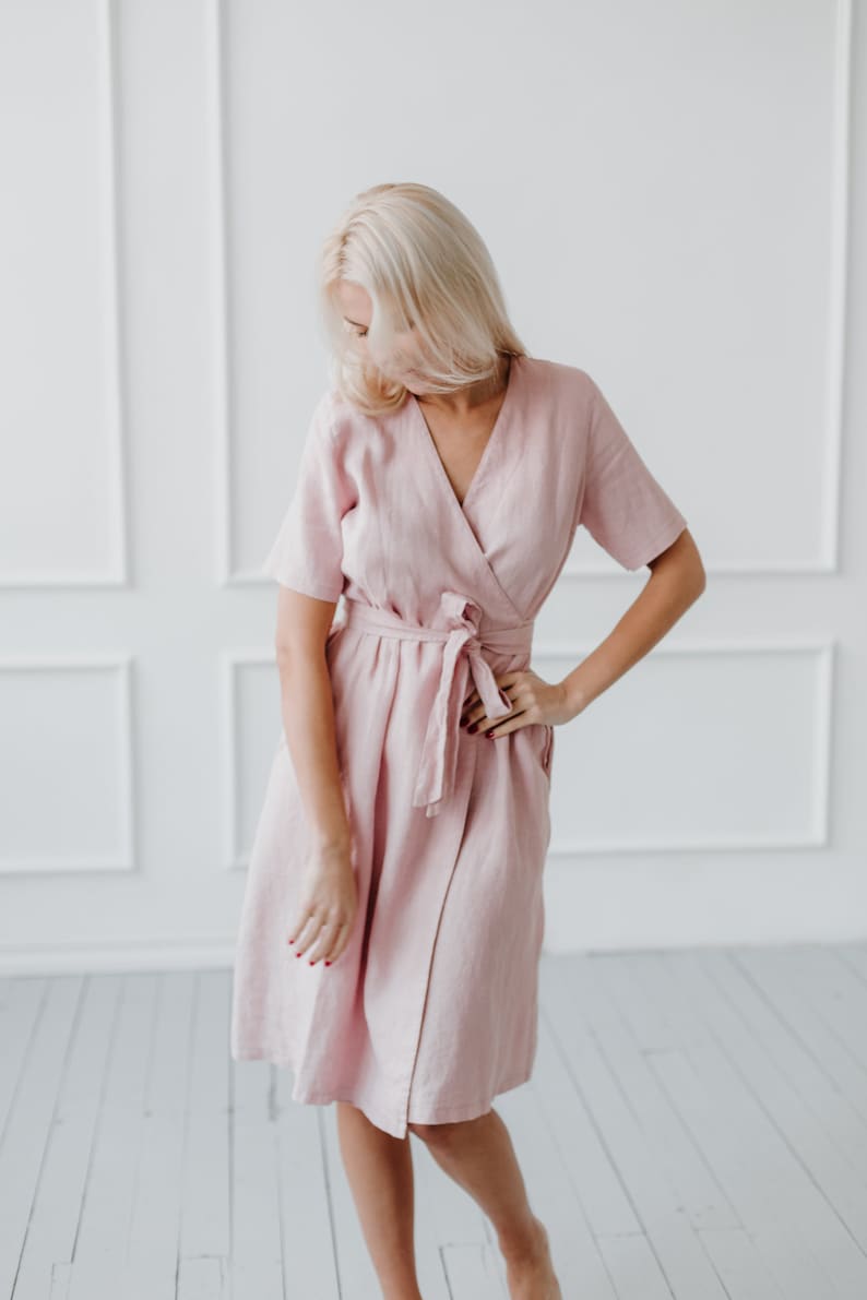 May include: A woman wearing a pink linen wrap dress with short sleeves and a tie at the waist.