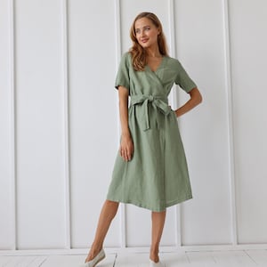 May include: A woman wearing a sage green linen wrap dress with a matching sash tied at the waist. She is standing on a white wood floor and wearing white canvas shoes.