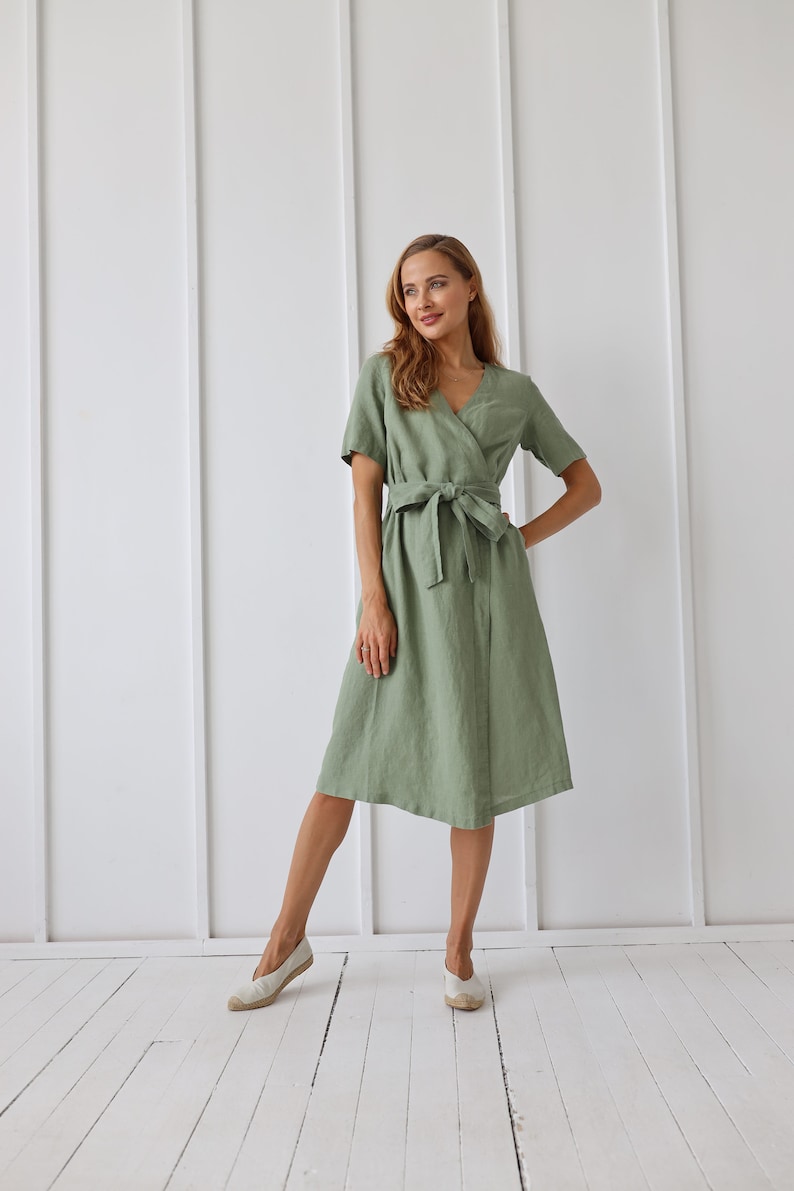 May include: A woman wearing a sage green linen wrap dress with a matching sash tied at the waist. She is standing on a white wood floor and wearing white canvas shoes.