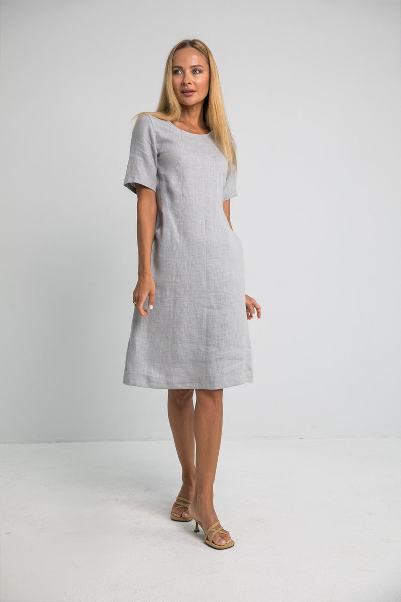 May include: A gray linen dress with short sleeves and a scoop neck. The dress is fitted through the bodice and flares out slightly at the hem. The model is wearing the dress with nude sandals.