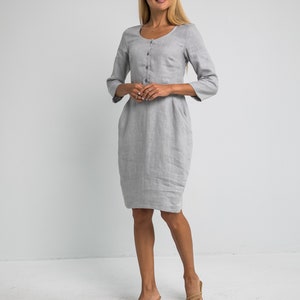 May include: A gray linen dress with a button-up front and three-quarter sleeves. The dress has a fitted bodice and a slightly flared skirt. The model is wearing the dress with nude heels.