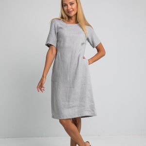 May include: A woman wearing a light gray linen dress with short sleeves and a scoop neck. She is standing with her hands in her pockets and is wearing tan sandals.