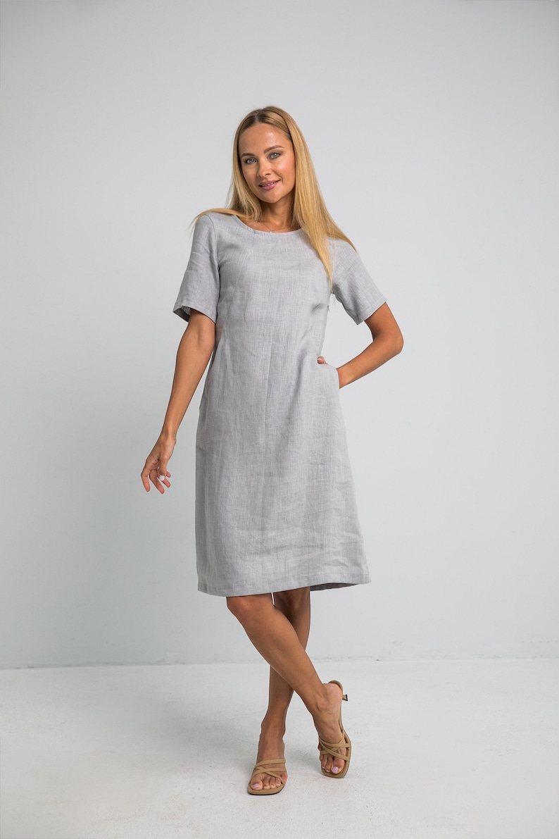 May include: A woman wearing a light gray linen dress with short sleeves and a scoop neck. She is standing with her hands in her pockets and is wearing tan sandals.