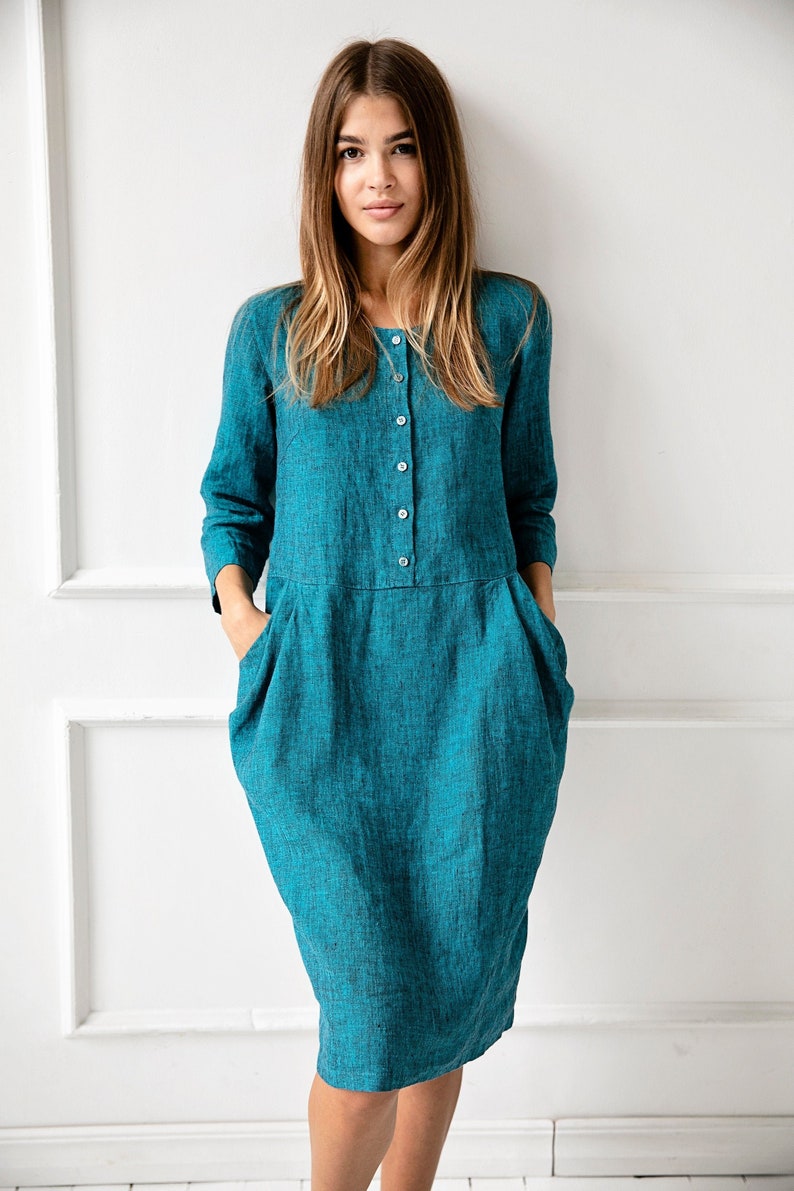 May include: A teal blue linen dress with a button-up front and pockets. The dress is knee-length and has a relaxed fit.