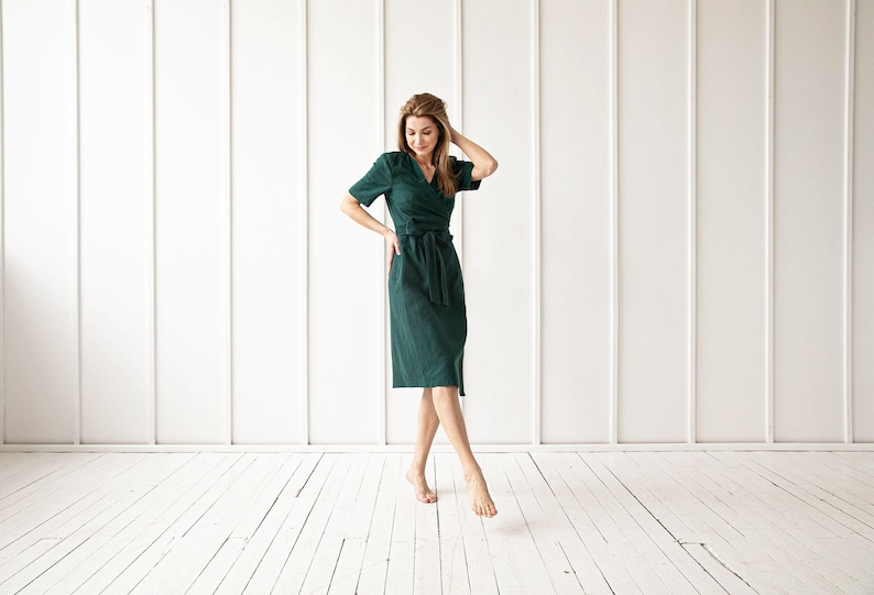 May include: A woman wearing a dark green wrap dress with a belt. She is standing on a white wood floor in front of a white wall.