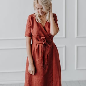 May include: A woman wearing a rust-colored linen wrap dress with a short sleeve and a tie belt.