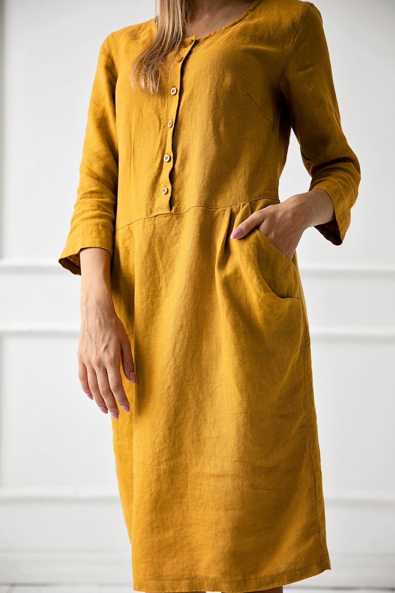 May include: A mustard yellow linen dress with a button-up front and pockets. The dress has a relaxed fit and a midi length.