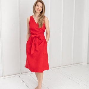 May include: A woman wearing a red sleeveless wrap dress with a belt. The dress has a V-neckline and pockets. She is standing on a white wooden floor.