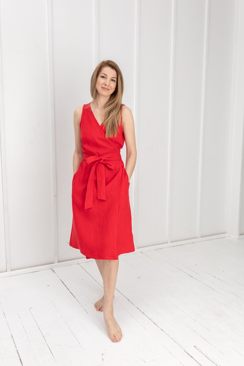 May include: A woman wearing a red sleeveless wrap dress with a belt. The dress has a V-neckline and pockets. She is standing on a white wooden floor.