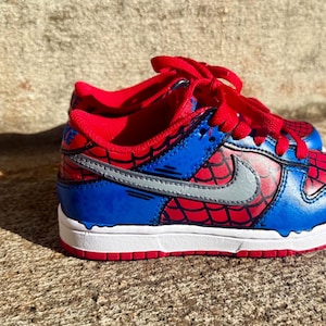 May include: A low-top sneaker featuring a vibrant red and blue color scheme with a black spiderweb pattern across the red panels. The shoe includes a silver Nike swoosh on the side, red laces, a white midsole, and a red outsole with blue accents.