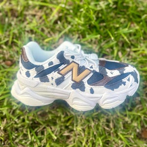 May include: White and blue athletic shoe with a cow print design. The shoe features a brown and gold logo on the side and white laces. The shoe is sitting on a bed of green grass.