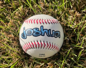 May include: A white baseball featuring red stitching and the name Joshua printed in a blue bubble-style font across the center. The bottom of the ball displays the text CHINA EHLC6 in small black letters, indicating its origin and production code.
