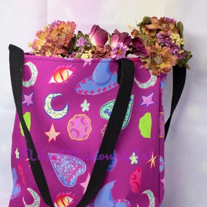 May include: A purple tote bag with black straps and a colorful print of moons, stars, hearts, and other magical symbols.