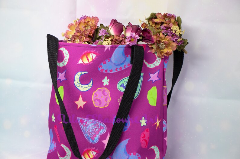 May include: A purple tote bag with black straps and a colorful print of moons, stars, hearts, and other magical symbols.