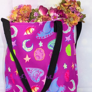 May include: A purple tote bag with black straps and a whimsical print of moons, stars, eyes, and witch hats. The bag is filled with a bouquet of pink and orange flowers.