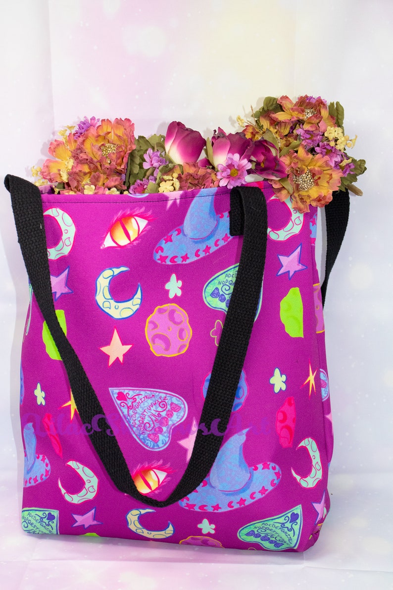 May include: A purple tote bag with black straps and a whimsical print of moons, stars, eyes, and witch hats. The bag is filled with a bouquet of pink and orange flowers.