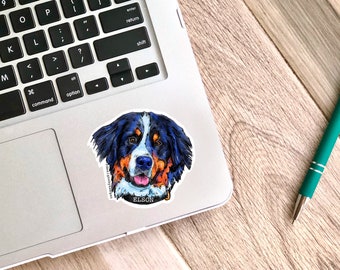 May include: A sticker of a Bernese Mountain Dog with blue, black, brown, and white fur. The dog is looking directly at the camera with its tongue sticking out. The sticker has the text "designsbykelgan" and "ELSON" on it.