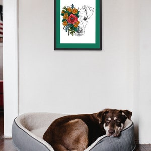May include: A framed print of a dog's head with flowers covering one eye. The dog is white and the flowers are red, orange, and yellow. The frame is black with a teal mat. A brown dog is lying in a gray dog bed.