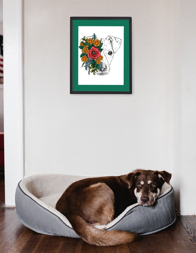 May include: A framed print of a dog's head with flowers covering one eye. The dog is white and the flowers are red, orange, and yellow. The frame is black with a teal mat. A brown dog is lying in a gray dog bed.