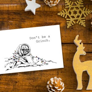 May include: A white card with black text that reads "Don't be a Grinch." A black and white illustration of a pug wrapped in a blanket with Christmas lights is on the card.