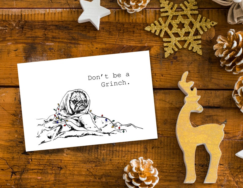 May include: A white card with black text that reads "Don't be a Grinch." A black and white illustration of a pug wrapped in a blanket with Christmas lights is on the card.