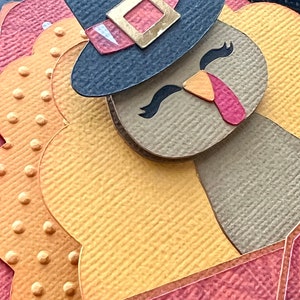 May include: A paper craft turkey with a pilgrim hat. The turkey features a brown body, yellow feathers, and a red wattle. The hat is navy blue with a gold buckle and a red band. The background is a mix of orange and red.