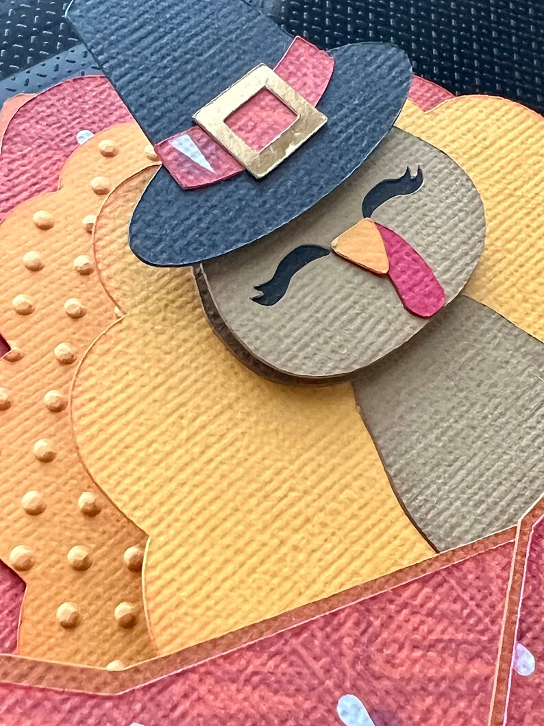 May include: A paper craft turkey with a pilgrim hat. The turkey features a brown body, yellow feathers, and a red wattle. The hat is navy blue with a gold buckle and a red band. The background is a mix of orange and red.